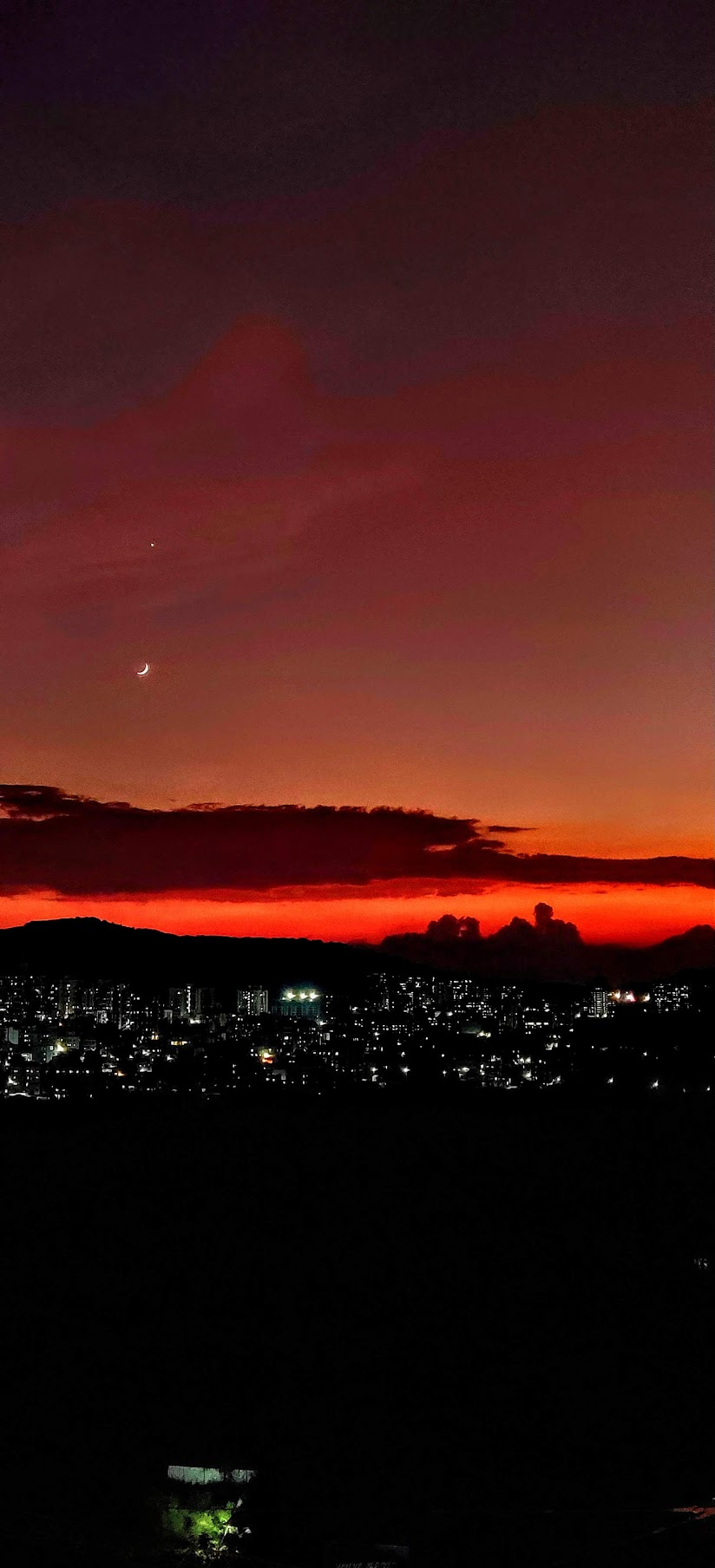 A crescent moon above a vivid red horizon and city lights
